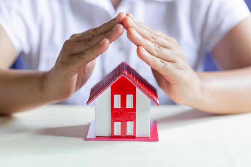 model małego domku z czerwonym dachem i złożone ręce człowieka nad nim